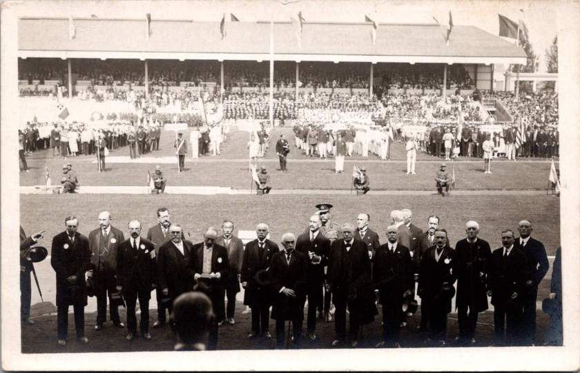 Sepia RPPC of military and civilian dignitaries gathered on a train platform between two carriages, with a row of soldiers in dress uniform standing at attention at right. Antwerp, 1920 Summer Olympics.